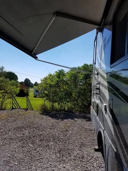 The view from a Heartland RV site out across the green field and treeline, awning extended on a sunny day